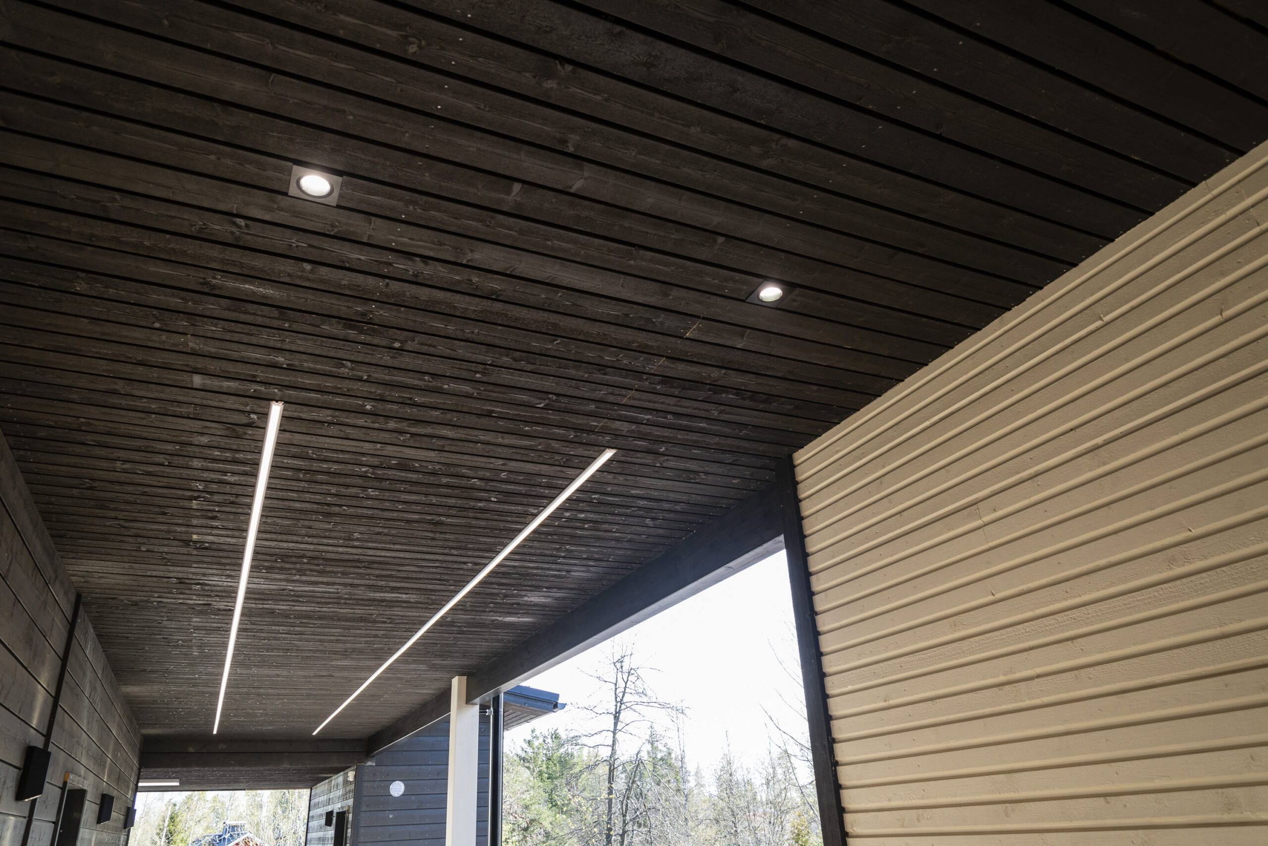 Moderni puinen katto, johon on upotettu LED-alasvalot ja tyylikkäät LED-linjavalot ulkokäytävän ylle. - LedStore Sisäänkäyntikäytävällä on kulkua valaisemassa kaksi pitkää led-nauhavaloa rinnakkain. Lisäksi kaksi uppoasennettua led-spottivalaisinta ja seinustalla lisäksi seinävalot.