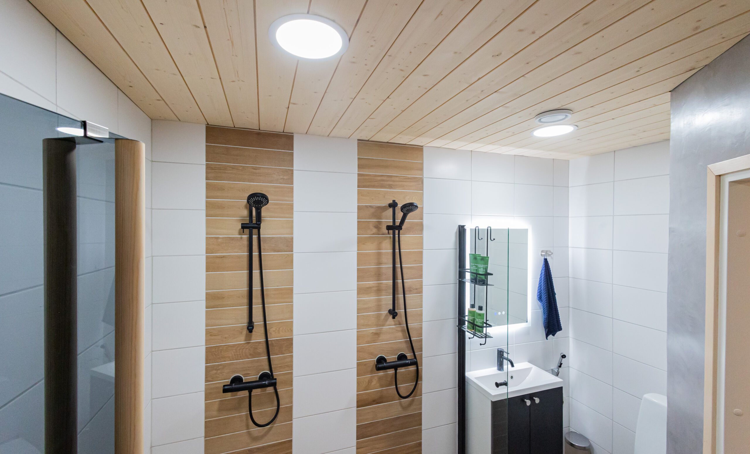Modernissa kylpyhuoneessa kaksi mustaa suihkua, tyylikkäät puupaneelit, valkoiset laatat sekä kompakti pesuallas peilillä. Tehosta tilan ilmettä LedStore.fi:n laadukkaalla led-valaistuksella! - LedStore Ledit kylpyhuoneessa, led-valopaneelit avaavat valoa laajalle pinta-alalle ja tuovat hyvän yleisvalon.