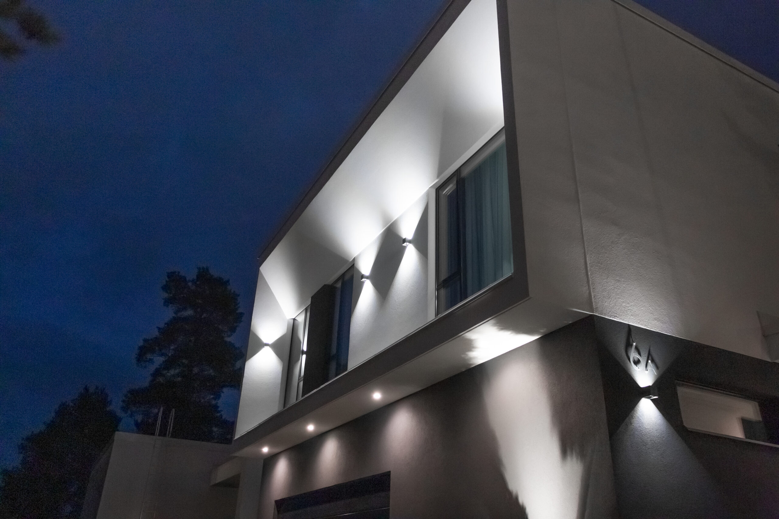 Moderni talon julkisivu yöllä, valkoiset seinät ja geometriset LedStore.fi:n led-valot valaisevat tyylikkäästi rakennuksen fasadin. - LedStore Led-pihavalaistus syntyy seinävaloista ja alaspäin osoittavista led-spoteista. Lisäksi seinävalaisin valaisee talon numeroa.