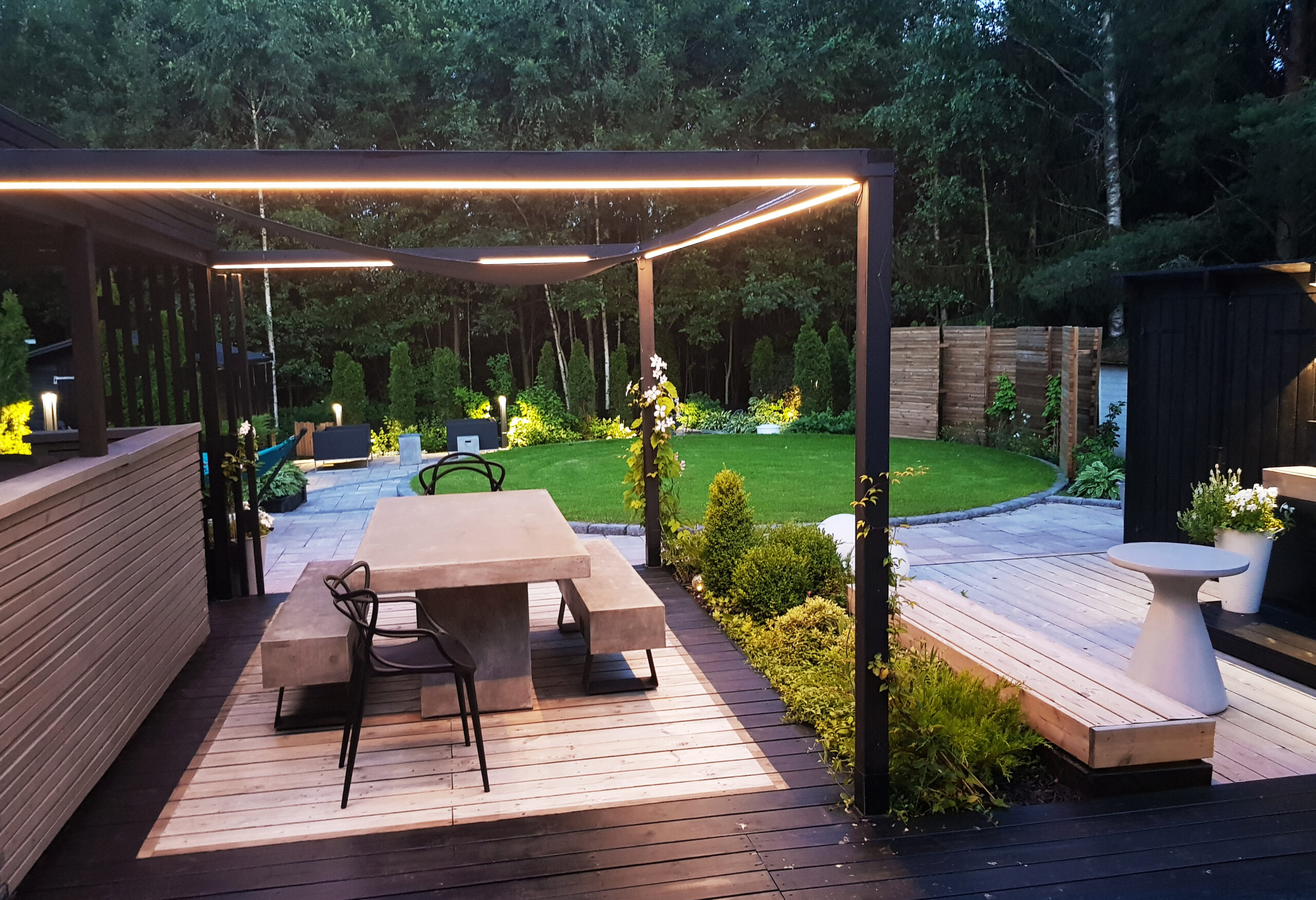 Moderni takapihan terassi, jossa on pergola, ulkoruokailuryhmä sekä vehreä nurmikko ja ympäröivät puut – valaistuksen kruunaa LedStore.fi:n tyylikäs led-valaistus. - LedStore Led-valaistus kesäkeittiöllä led-nauhavaloa katoksen kehikossa