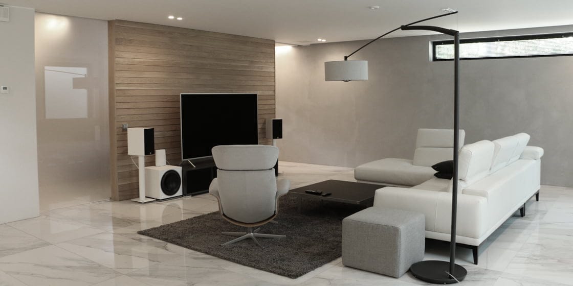 Modern living room with LED-backlit TV, white sectional sofa, gray armchair, black floor lamp, marble floor, and wooden accent wall with speakers.